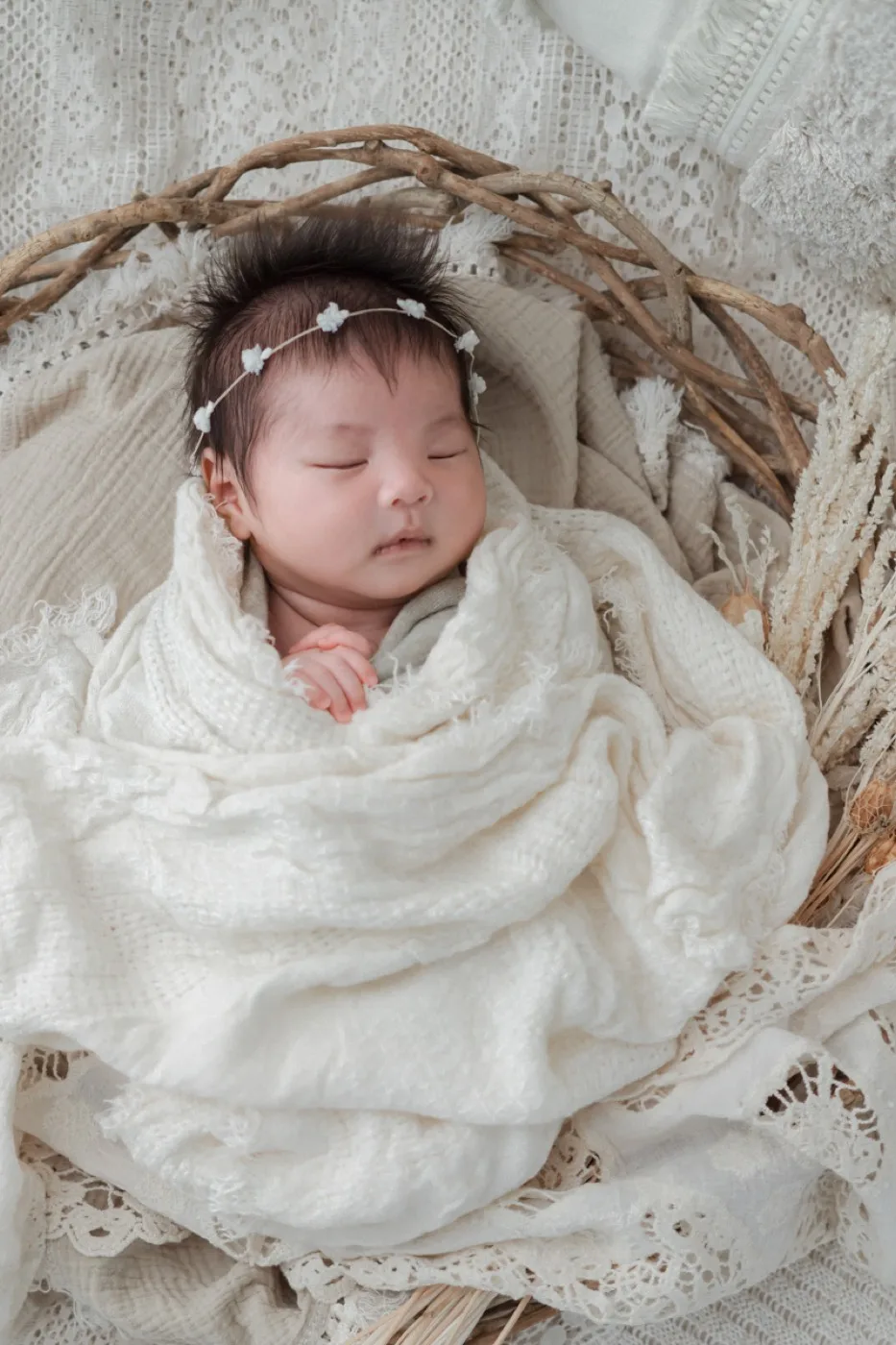 かごの中で白いおくるみに包まれ、花冠をつけて眠る新生児