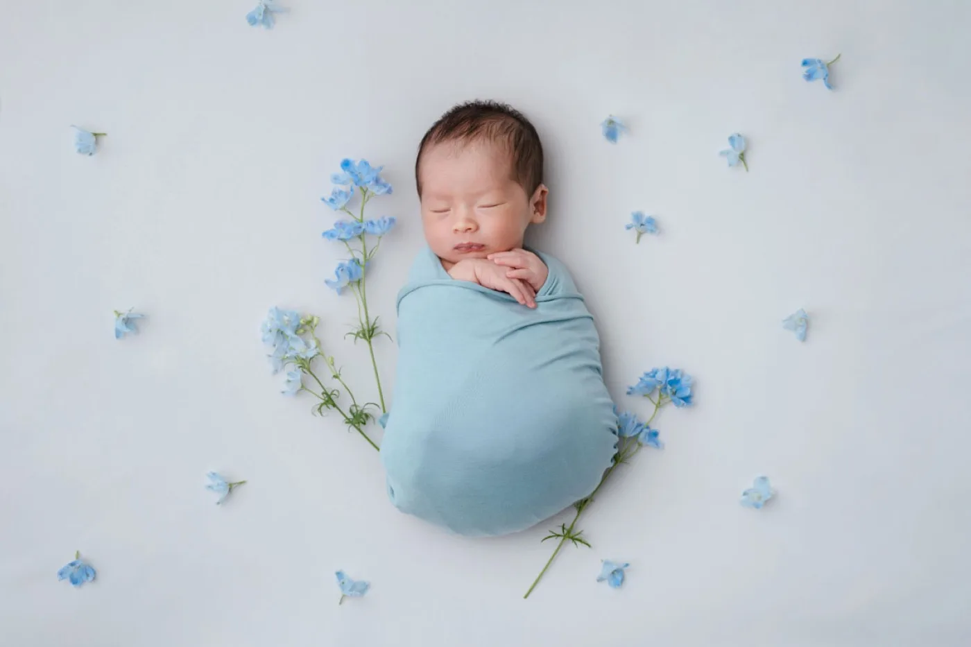 白い背景に青い小花を散らし、水色のおくるみで眠る新生児