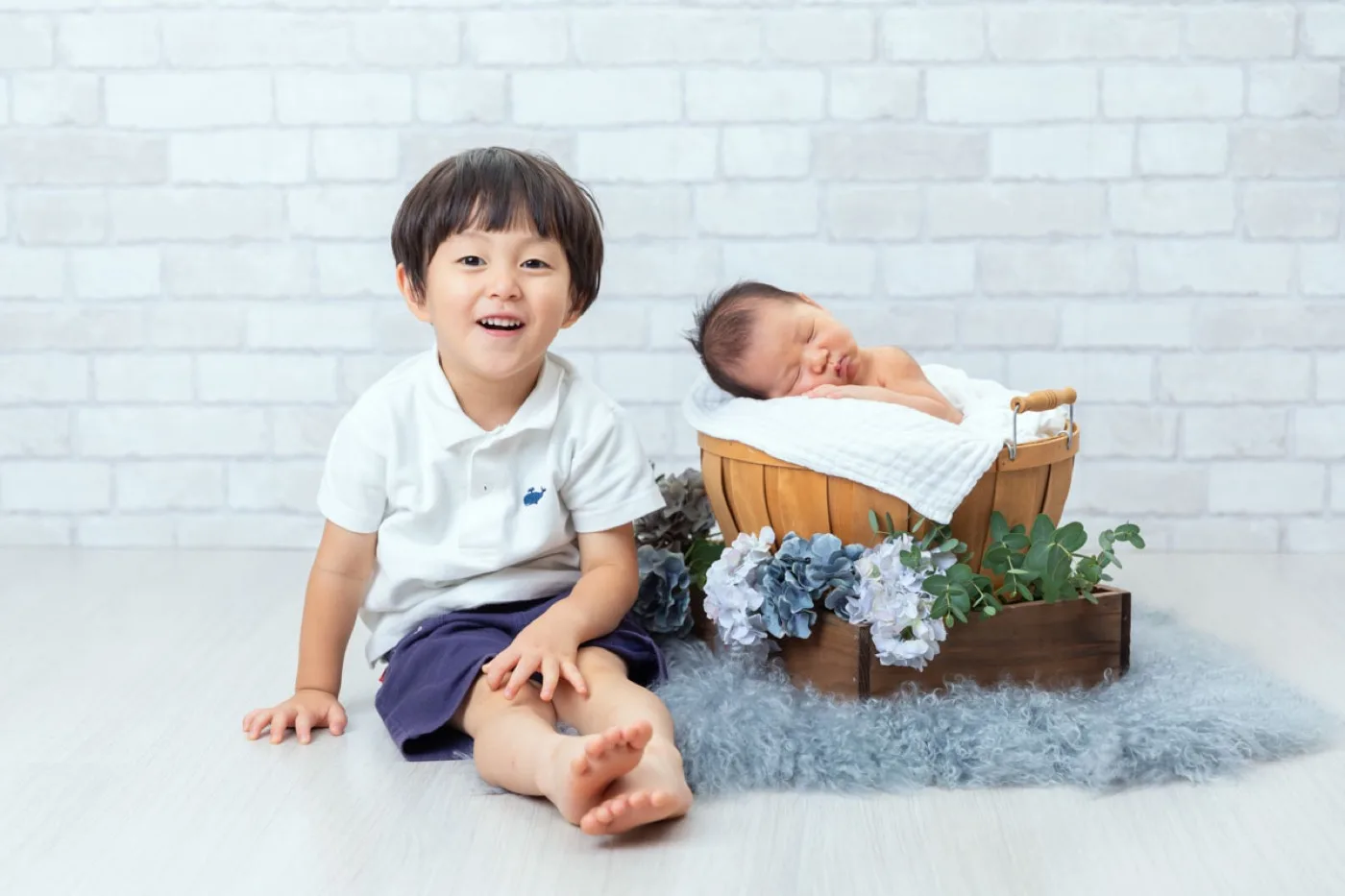 レンガの壁の前で、かごに眠る新生児のとなりに座って笑うお兄ちゃん