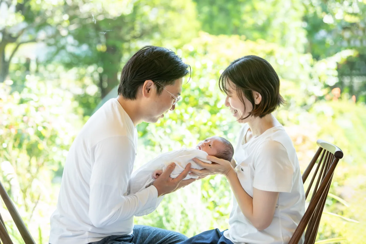 庭の緑を背に、おくるみに包んだ赤ちゃんを両親で見つめる