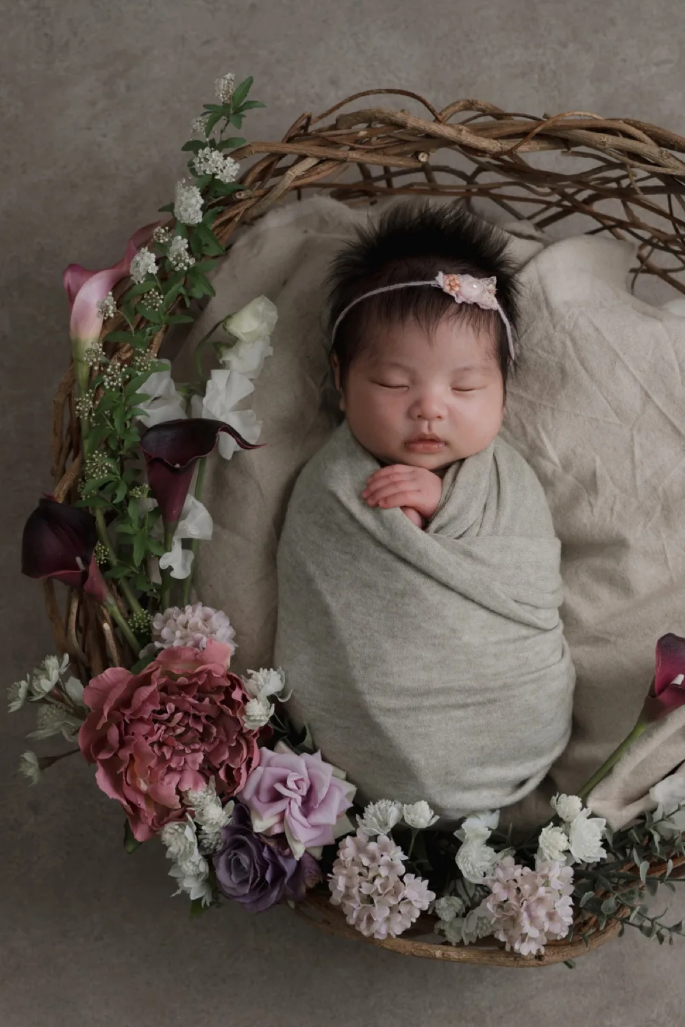 花のリースを添えたかごの中で、生成りのおくるみに包まれて眠る新生児