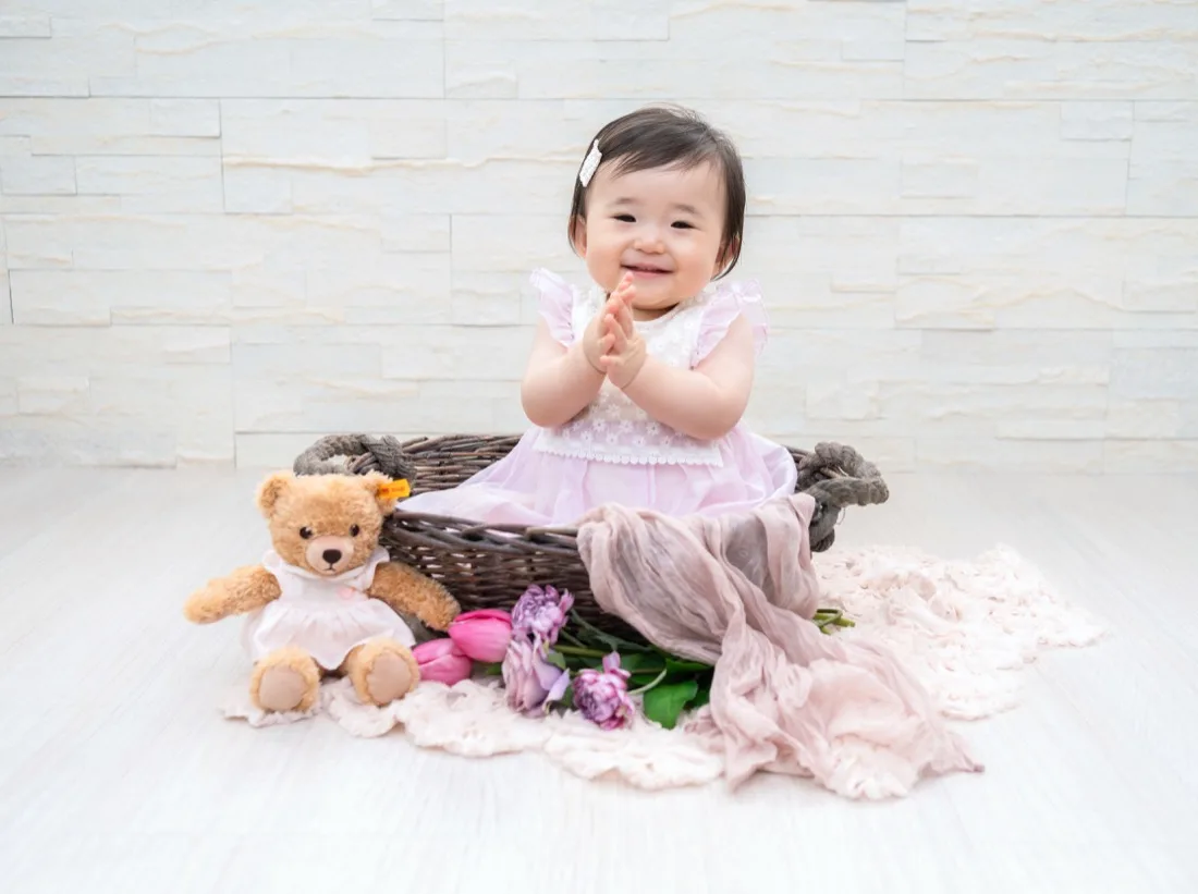 かごの前に座って手を合わせる1歳の子どもと、となりのテディベア