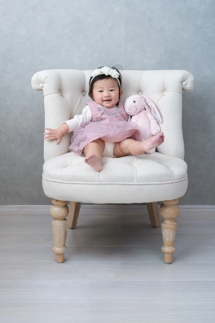 白い椅子に座り、うさぎのぬいぐるみを抱く生後6か月の赤ちゃん